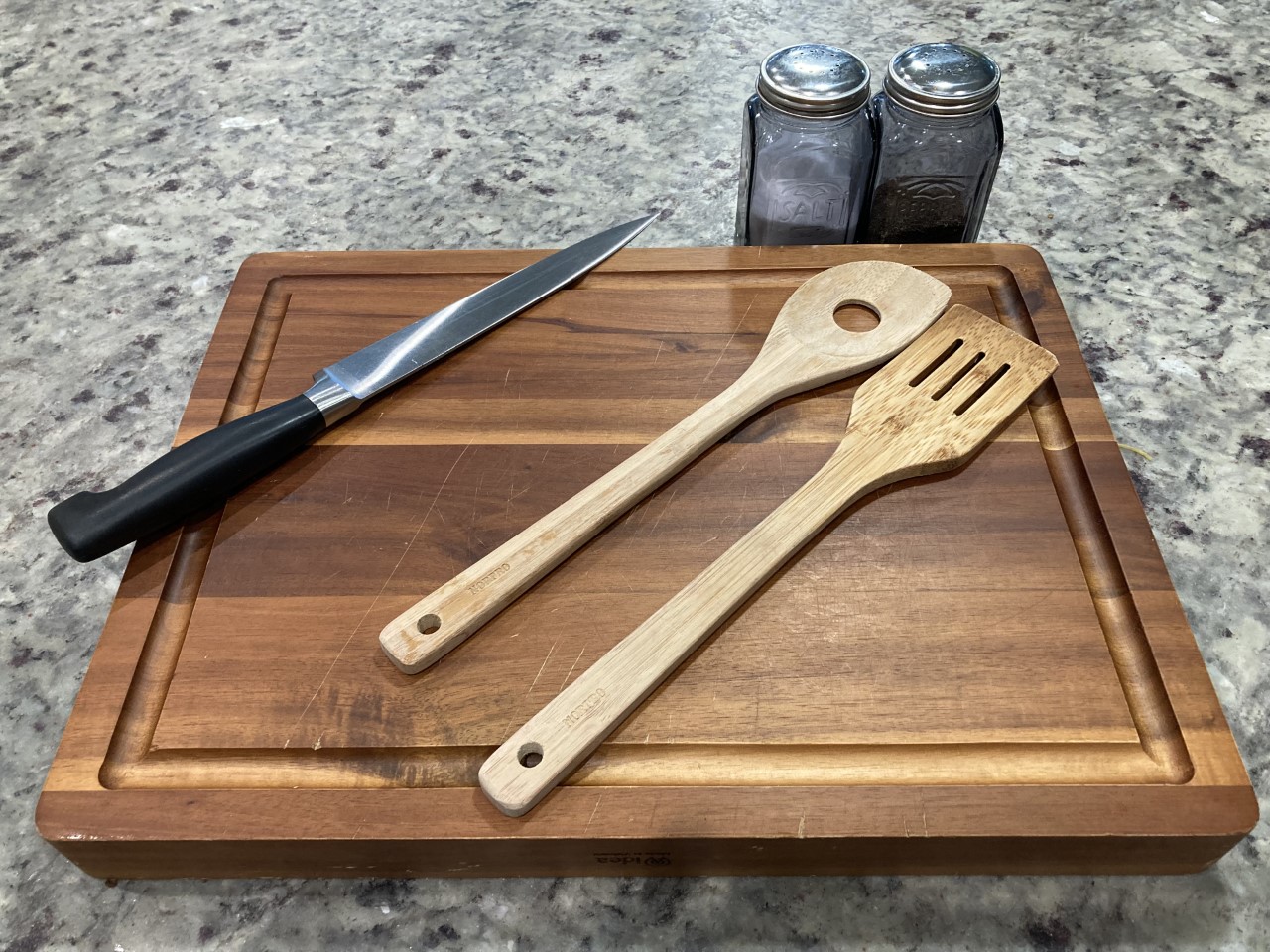 Kitchen cutting board with a knife, salad tongs, and salt and pepper shaker next to it.